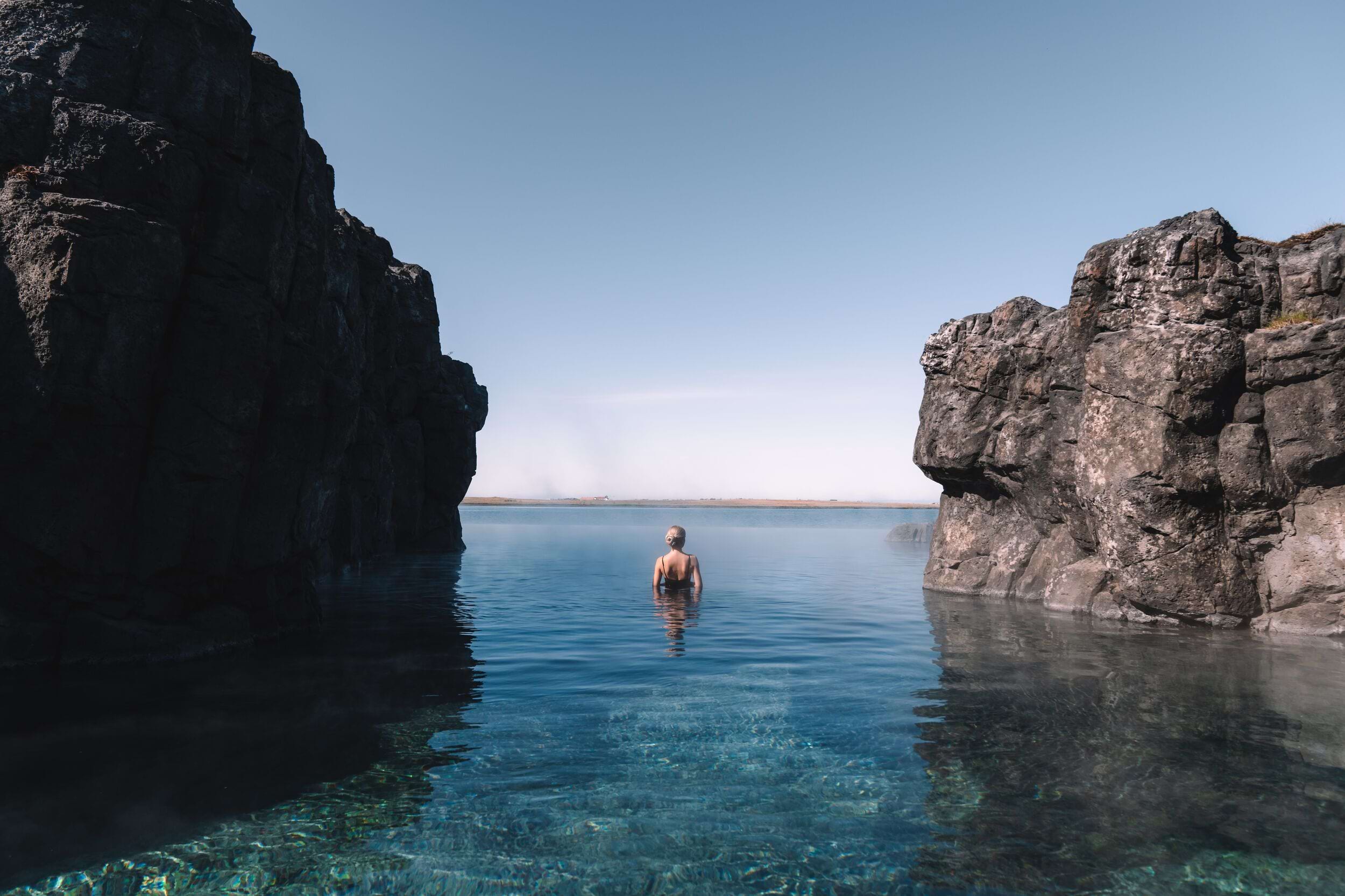 A single swimmer in the Sky Lagoon with blue skies and the rising sun in the background